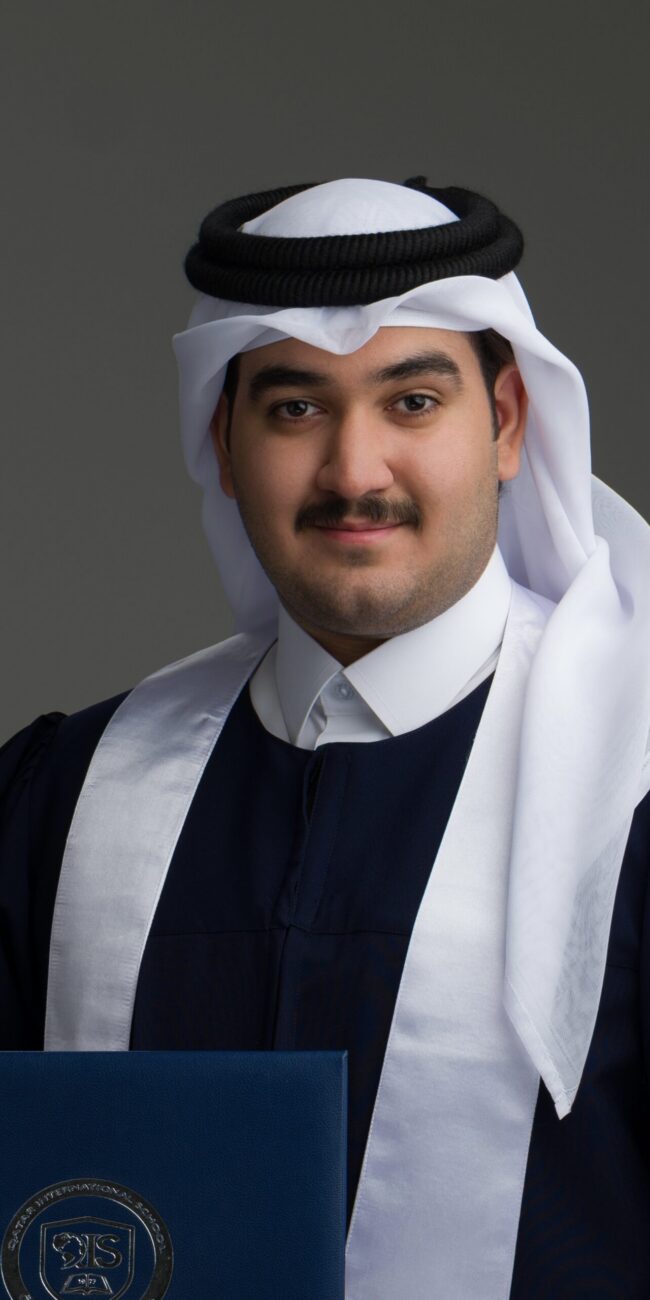 Studio graduation portrait of a young Qatari man in ghutra and agal, wearing a navy gown and holding his diploma folder.